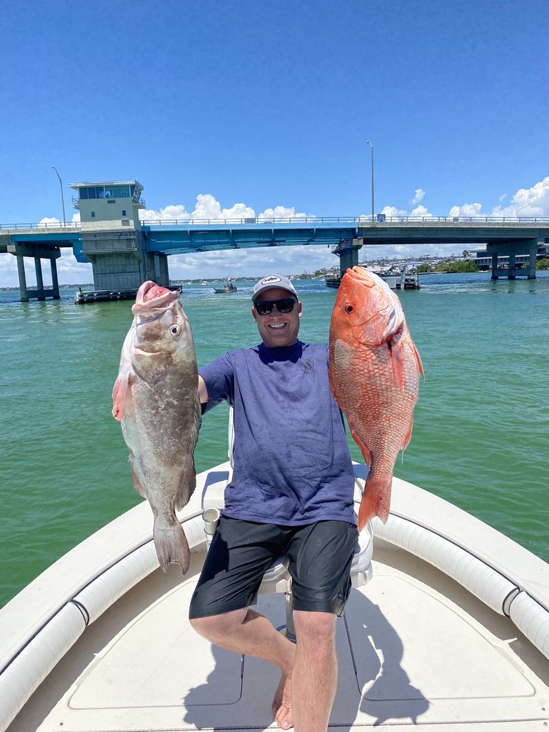 An angler on the bow holding a gag grouper in one hand and a red snapper in the other, a bascule bridge behind