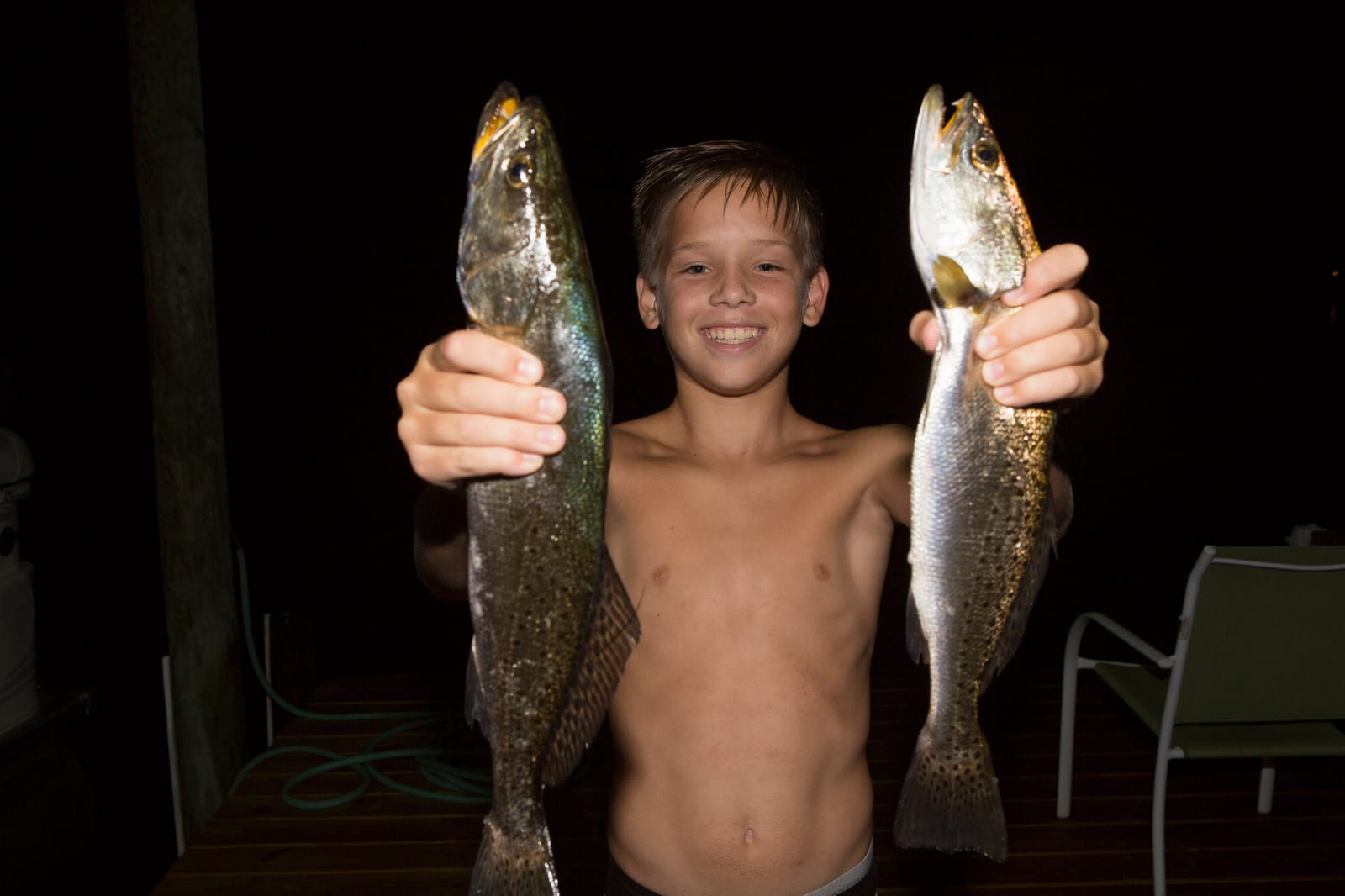A boy holding up two spotted seatrout, one in each hand, on a dock at night